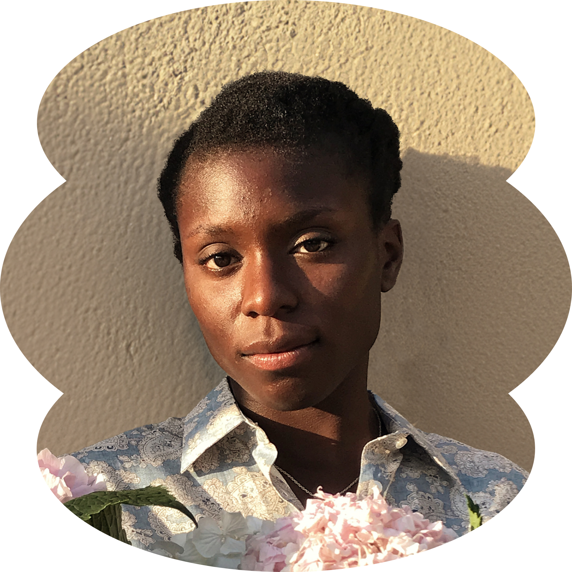 Black female holding flowers and standing against a beige wall in the sunlight